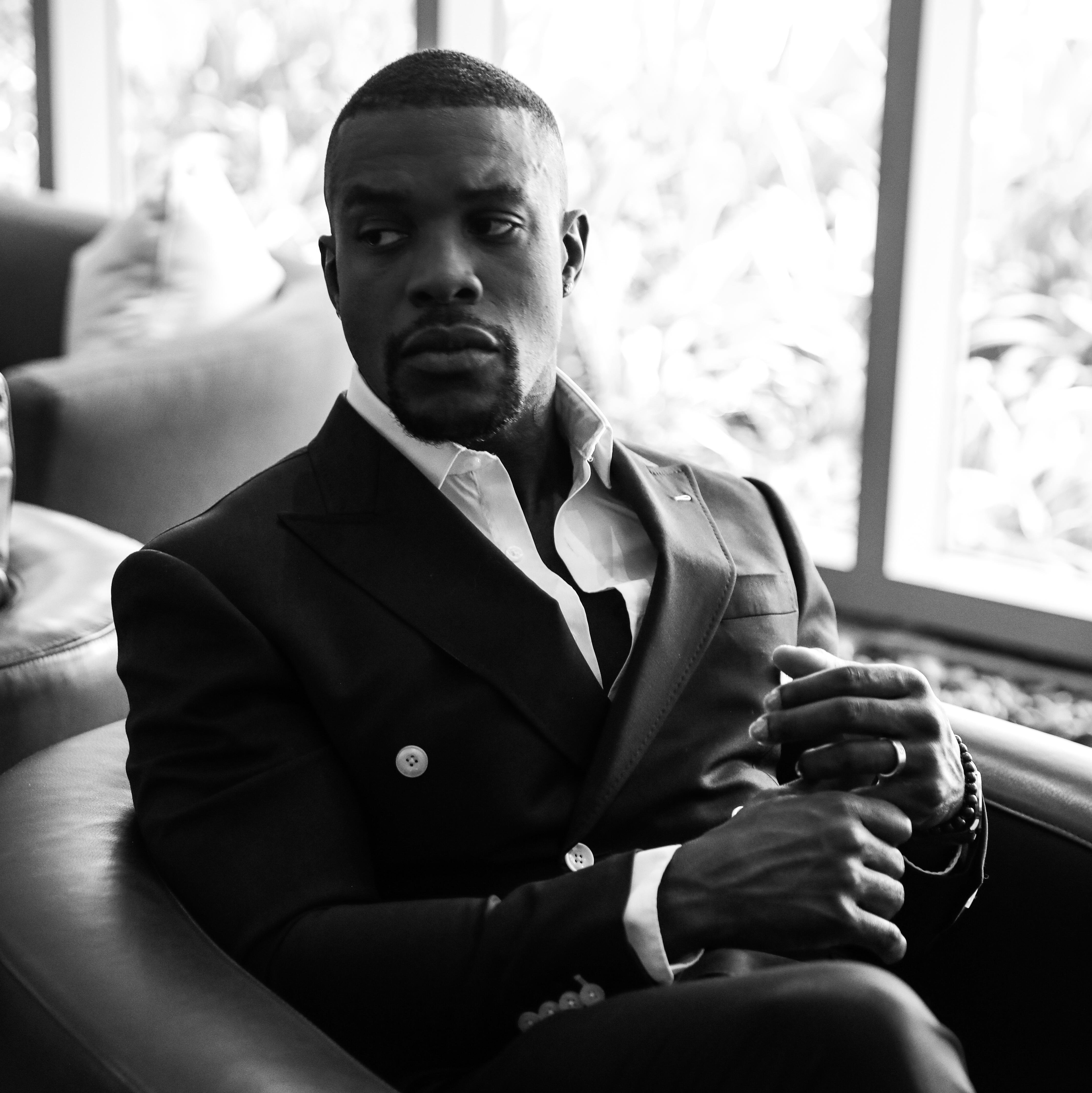 Patrick Ductant wearing his trademark suit sitting in a chair by a window.